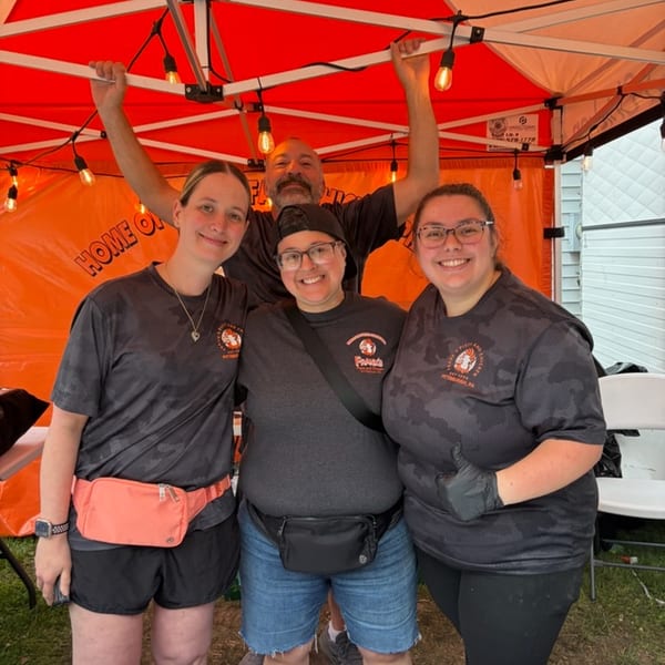 Four smiling people in branded shirts pose under an orange tent with string lights.