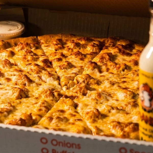 A Buffalo Chicken pizza in a box with a bottle of sauce and dipping cups.